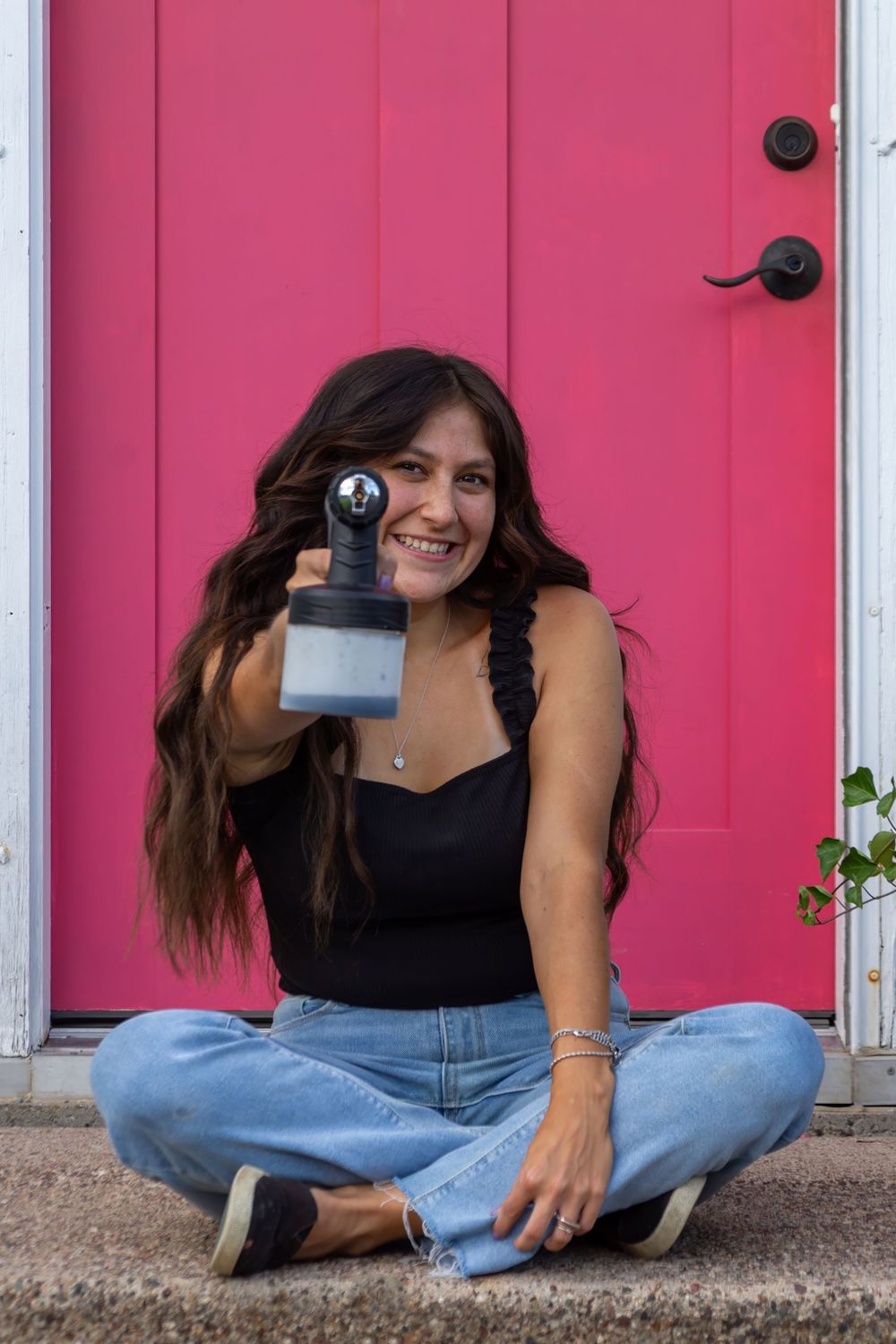 Person sits cross-legged in front of bright pink door holding paint sprayer while wearing black top and jeans.