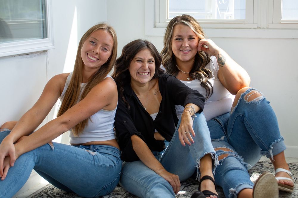 Three friends wearing casual outfits sit together smiling and relaxing on a couch by a window.