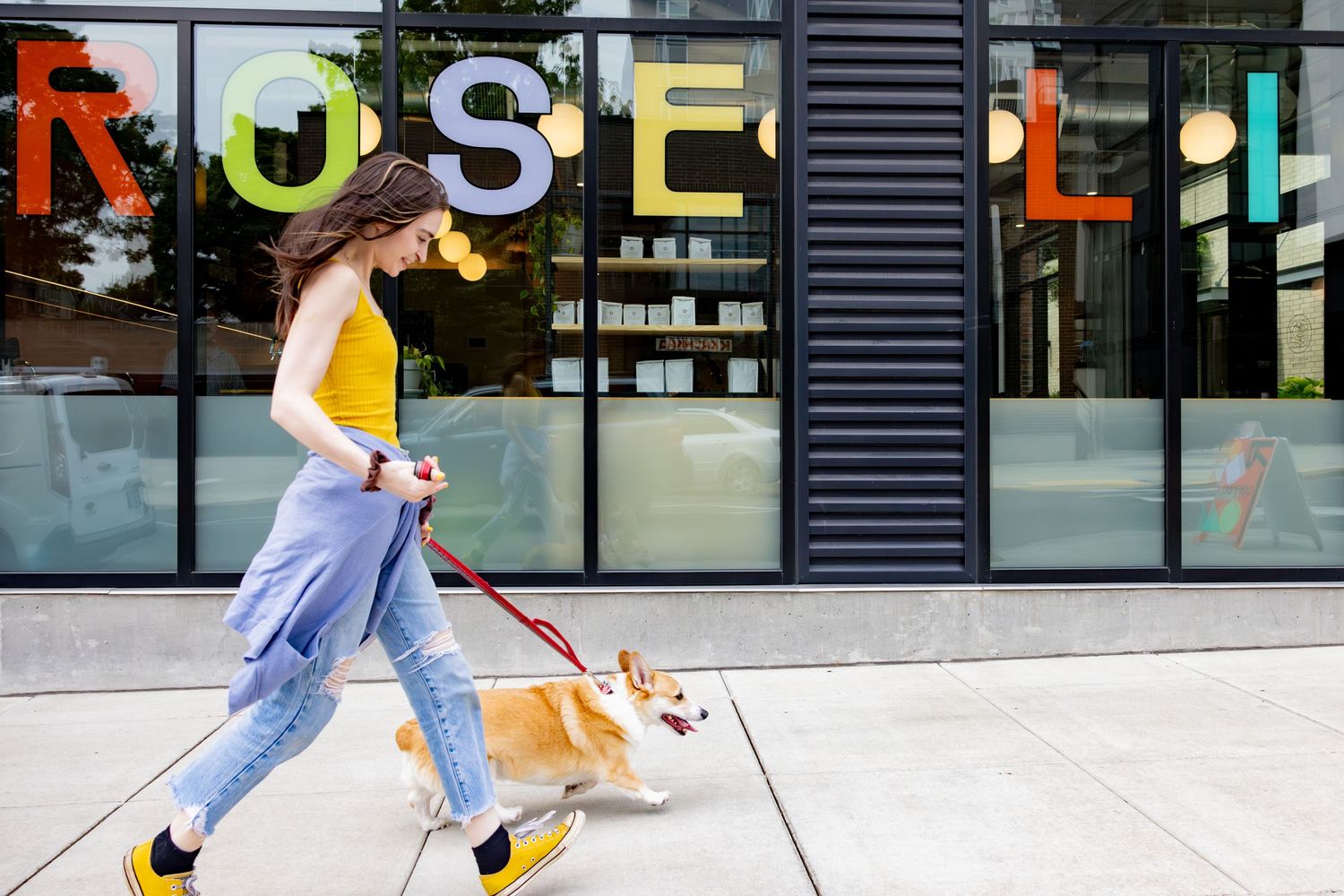 Someone in a yellow tank top and blue jeans walks a small corgi dog past a storefront with colorful signage.