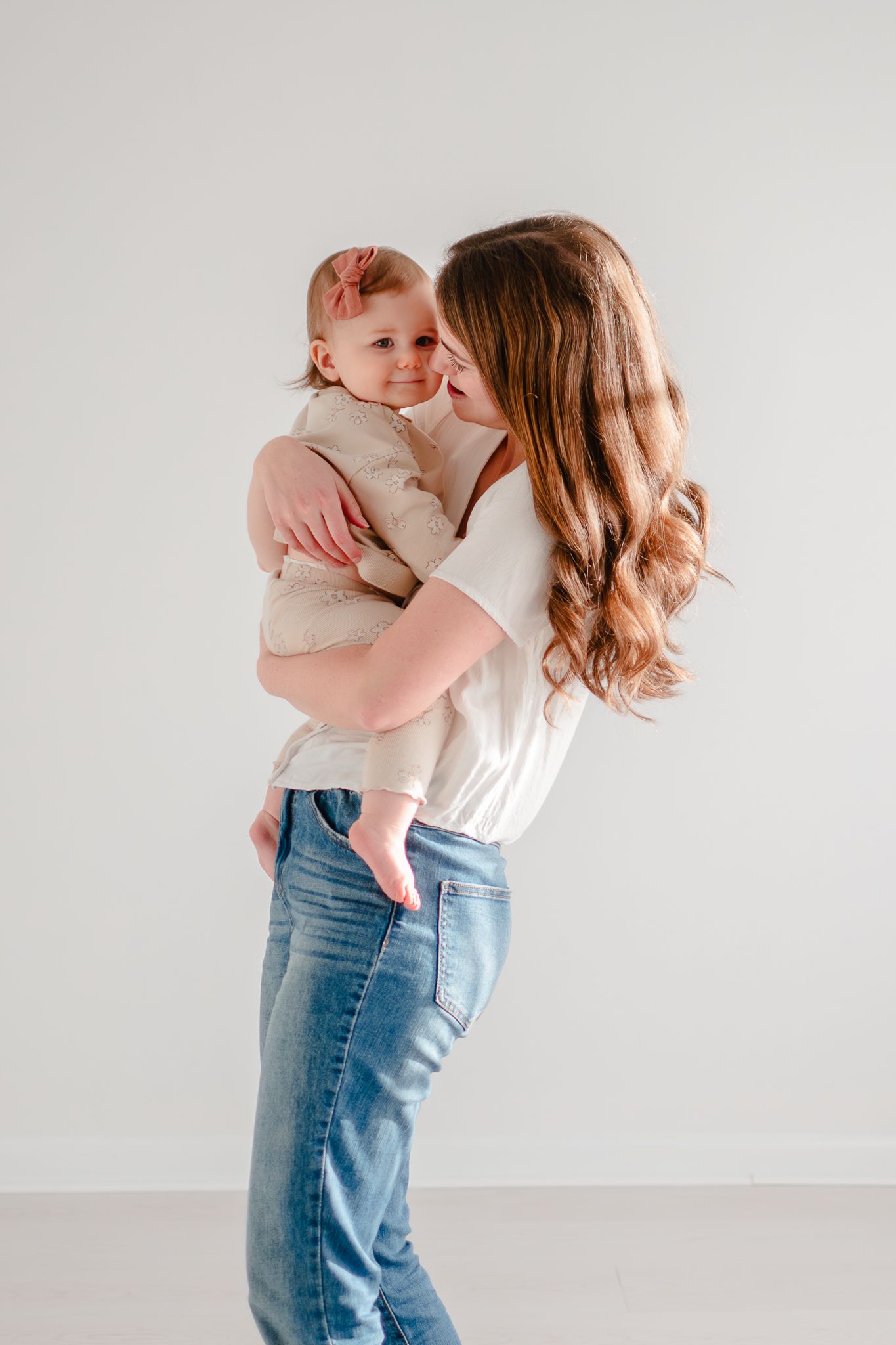A mother in jeans and white shirt holds a baby wearing a cream-colored outfit against a light background.