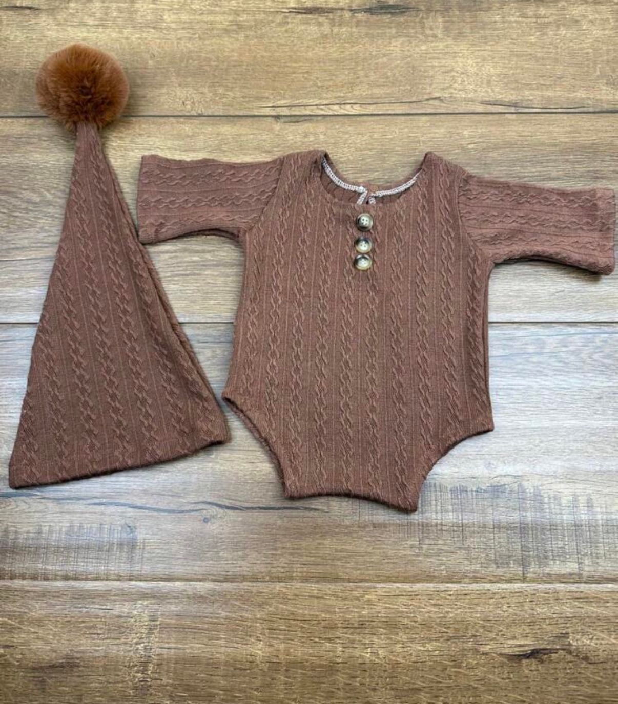 Brown knit baby romper and matching hat with pom pom on wooden background.