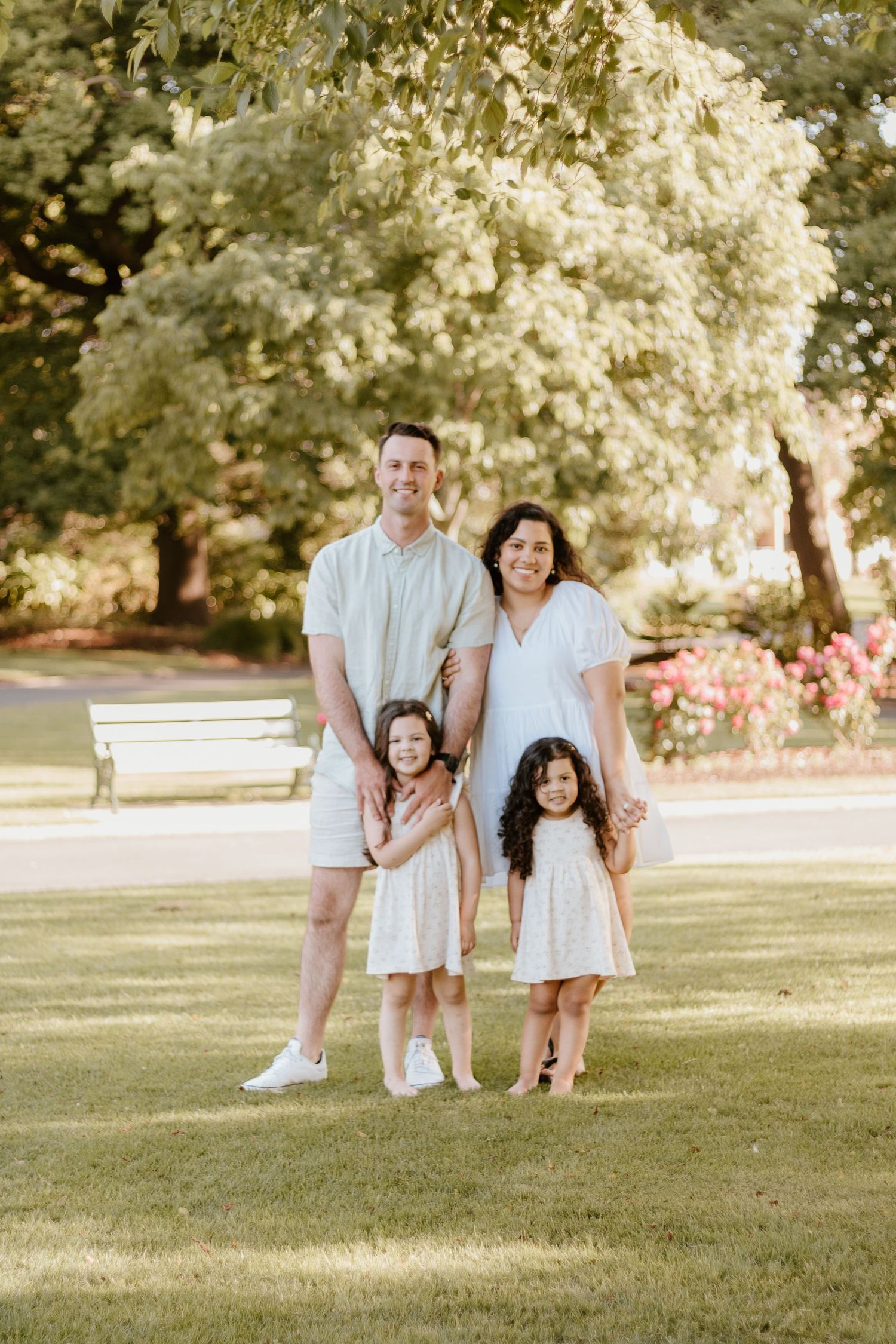 Family of 4, mum, dad, two young girls, standing together at a Launceston park smiling
