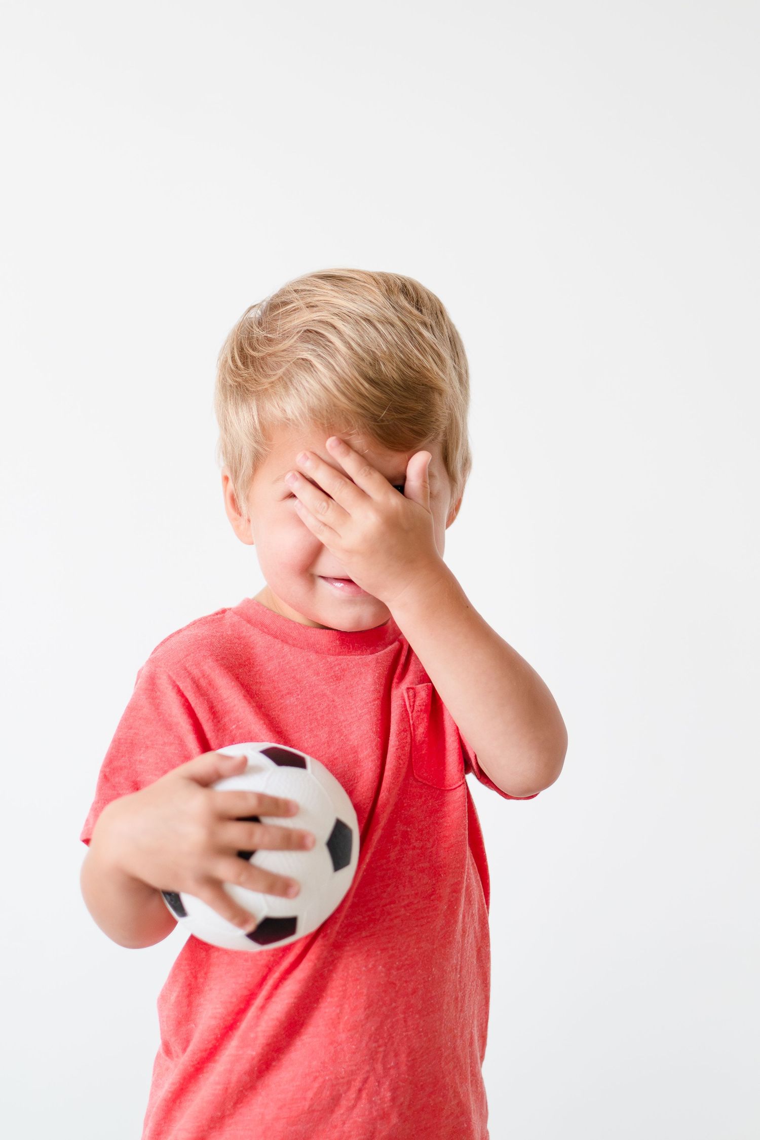 Child in coral red t-shirt covers face with hand while holding small soccer ball against white background.