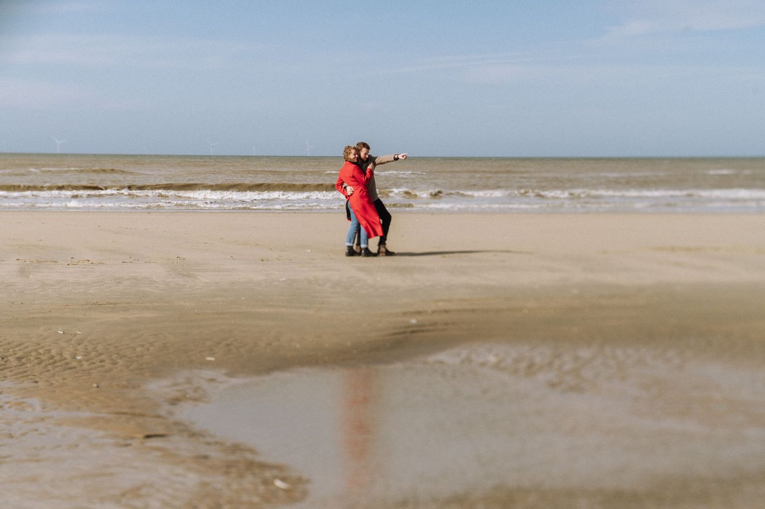 Two figures in red embrace on a vast empty beach during low tide.