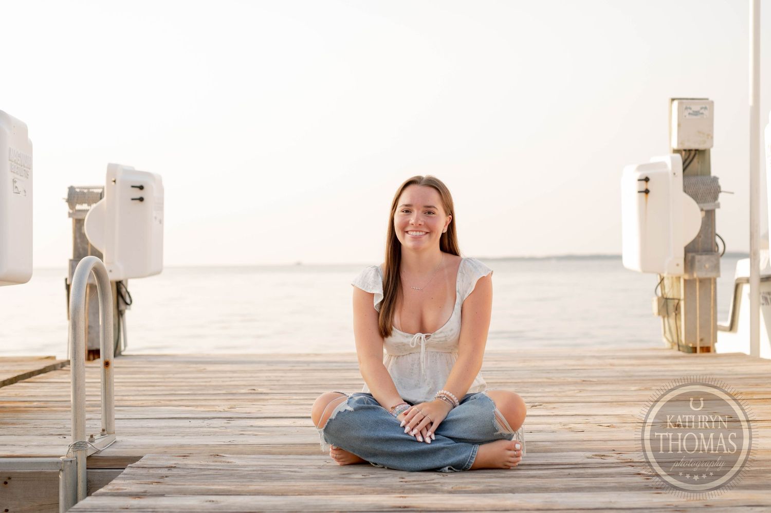 Person in white top and jeans sitting cross-legged on waterfront dock at dusk.