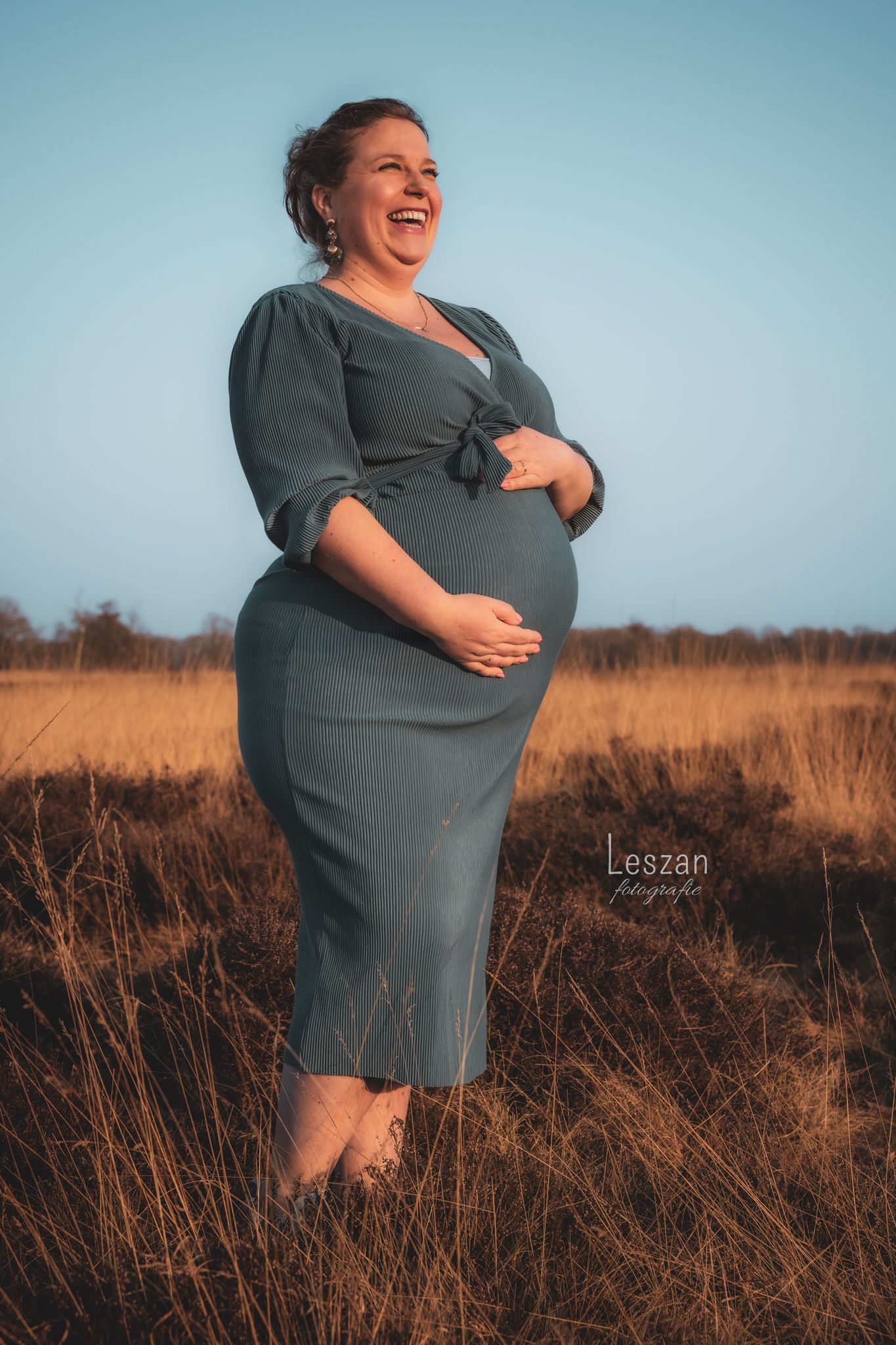 Pregnant person wearing a gray dress stands joyfully in a golden field at sunset against blue sky.