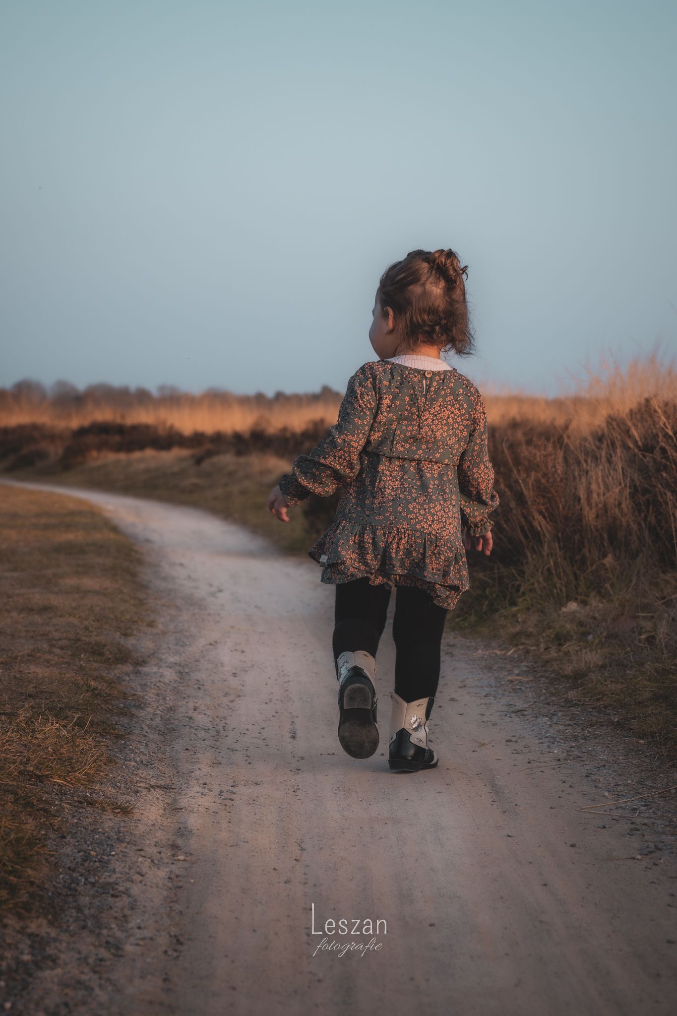Child wearing a floral dress walks alone on a dusty rural path during golden hour.