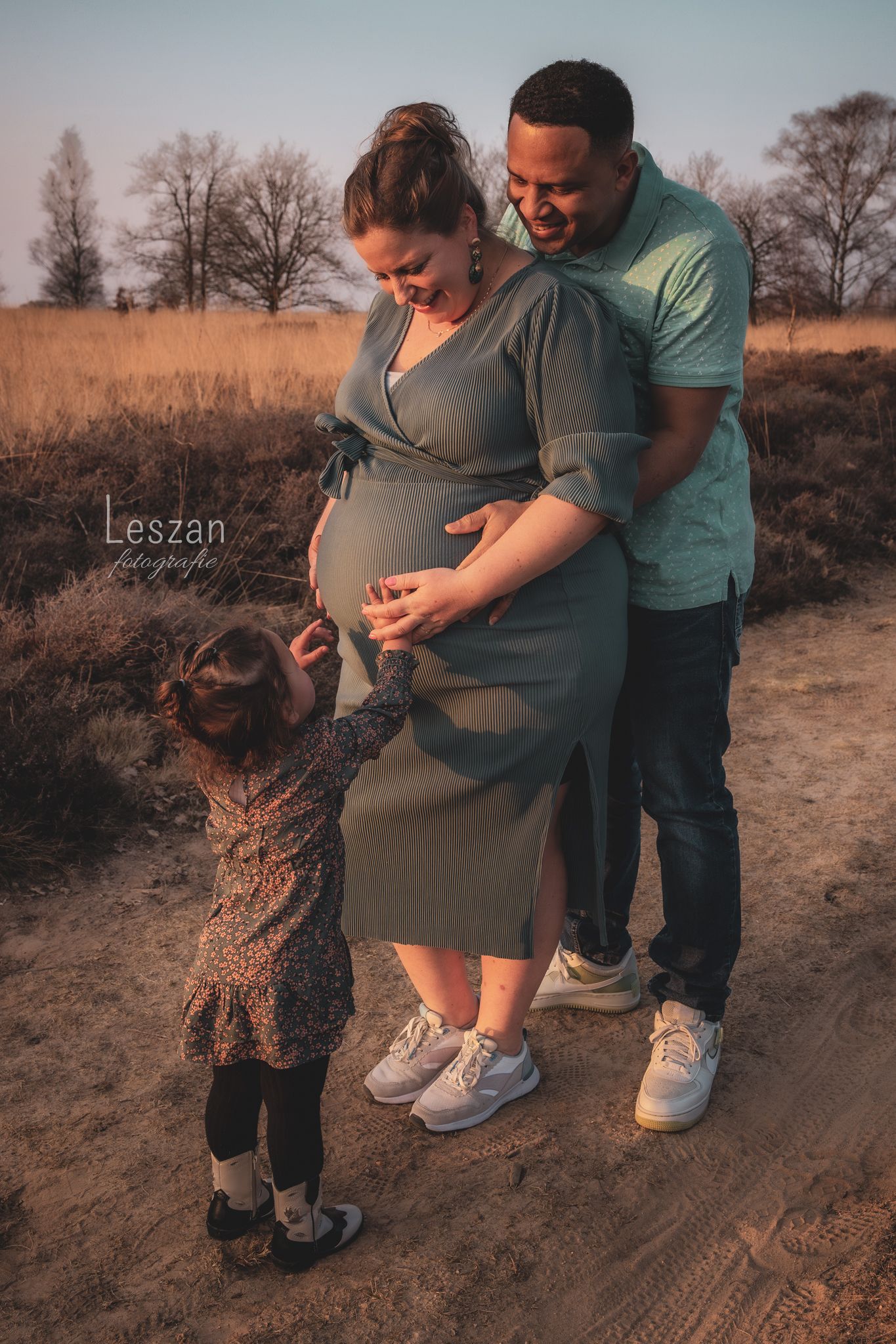 A couple and small child share a tender moment during an outdoor maternity photoshoot in a rustic field at sunset.