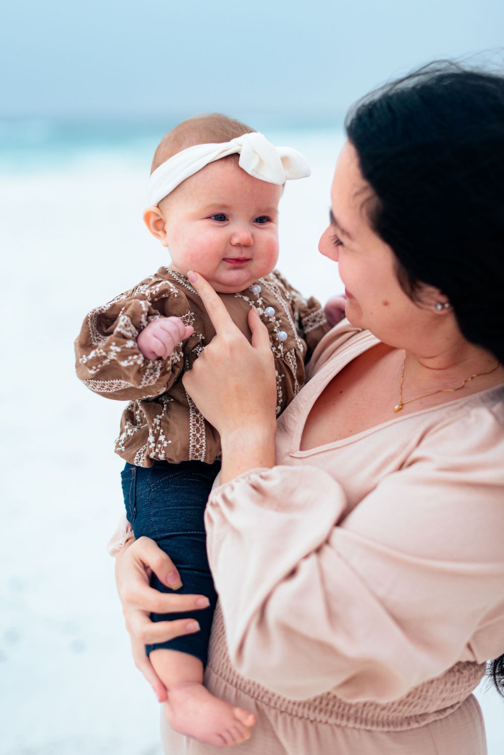 A sweet interaction between parent and baby wearing a white bow headband at the beach.