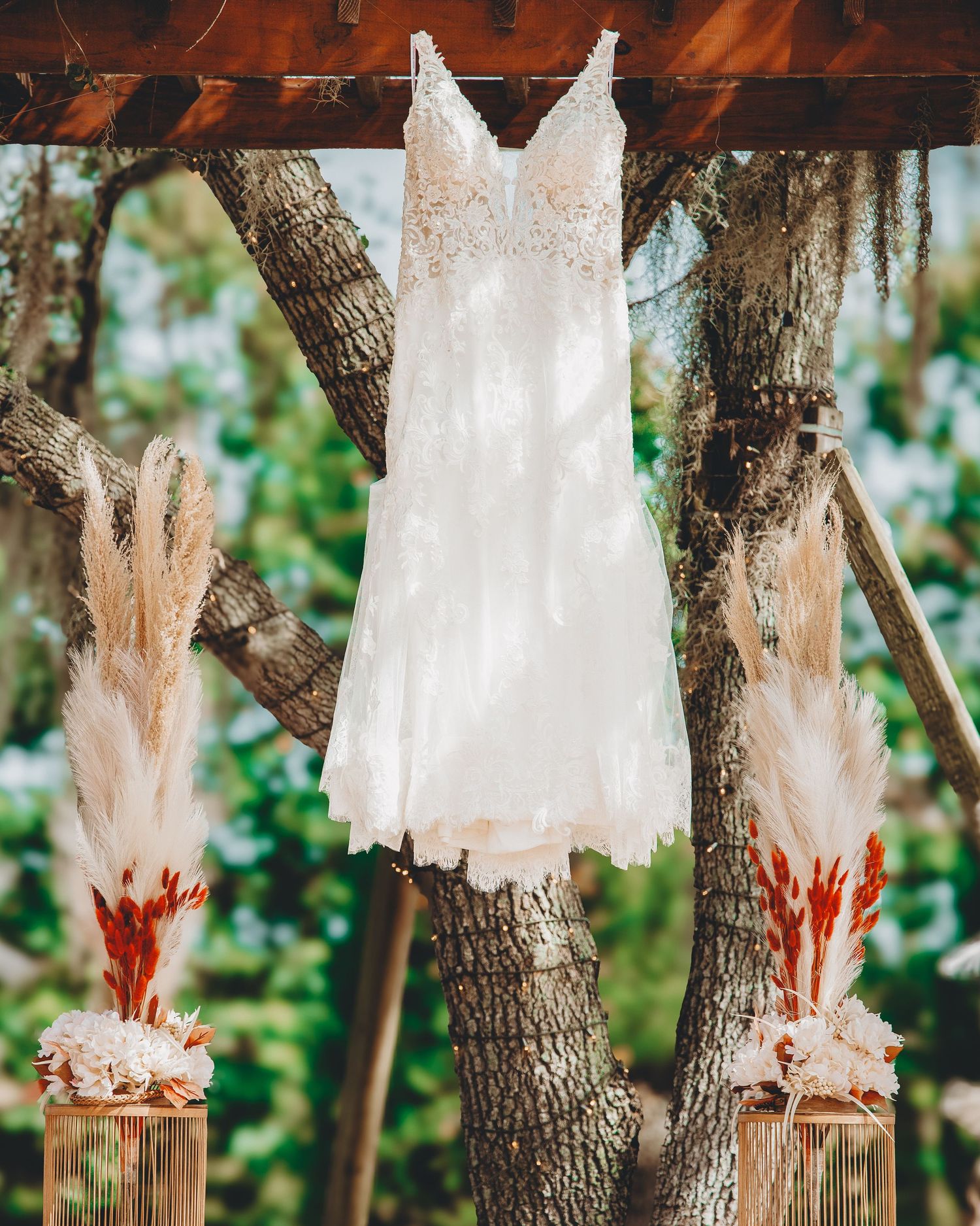 A white lace wedding dress hangs on a tree branch with pampas grass floral arrangements on either side.