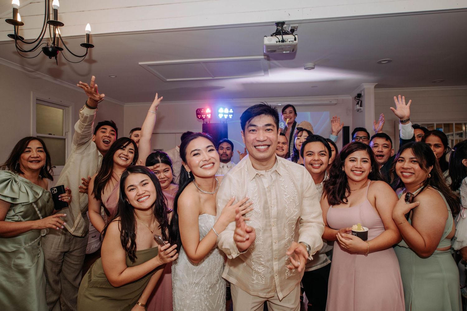 Wedding reception celebration with guests in pastel attire dancing and cheering in an indoor venue.