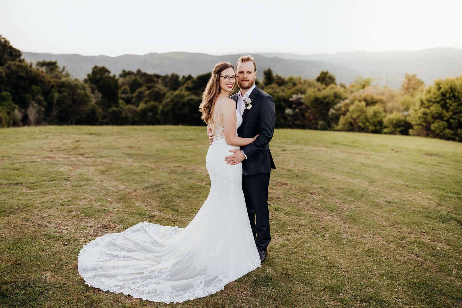 A wedding couple poses together on a hilltop meadow with mountains in the background during golden hour.