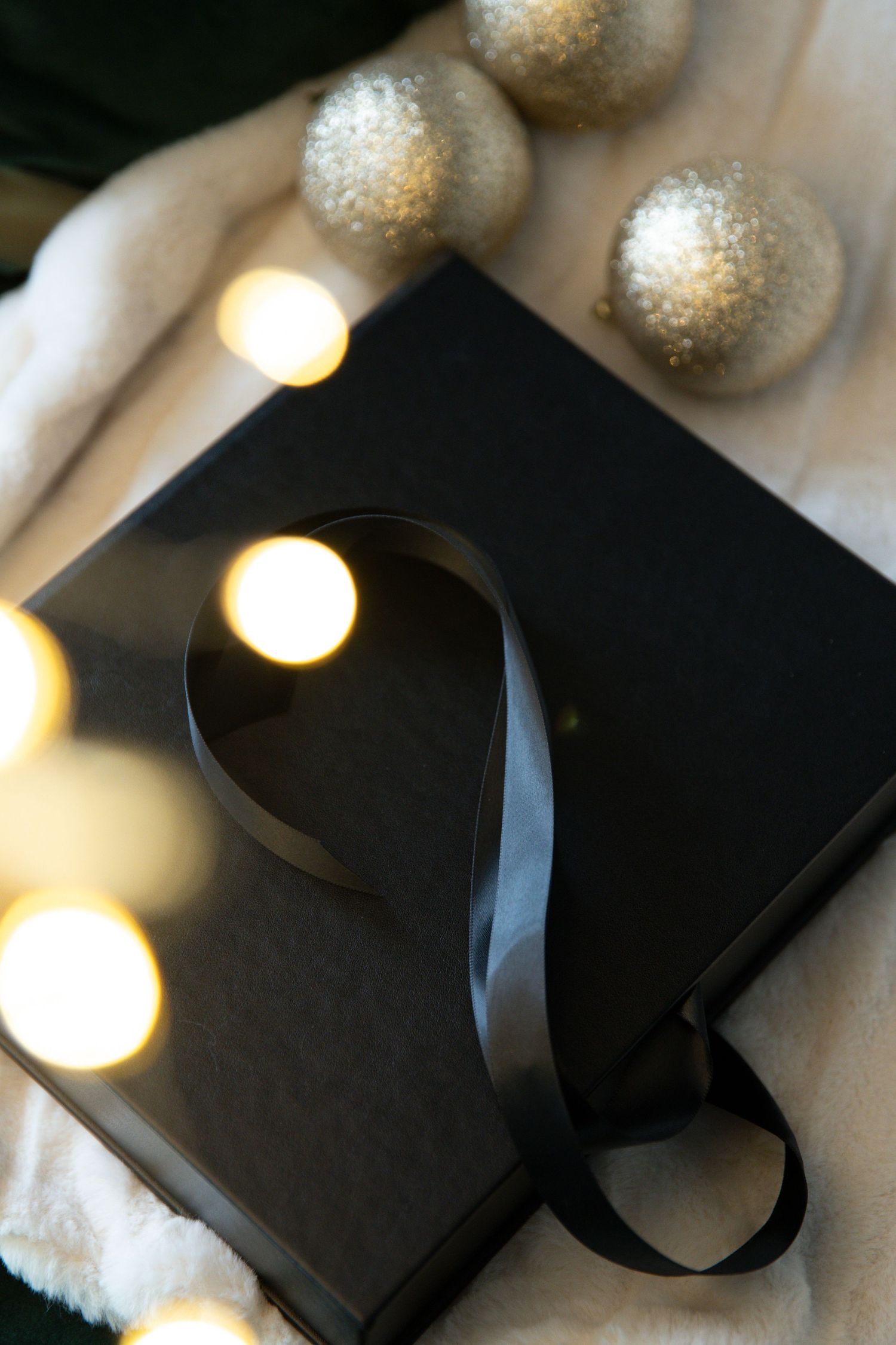 A black gift box with ribbon sits on white fabric next to glittery silver ornaments and glowing lights.