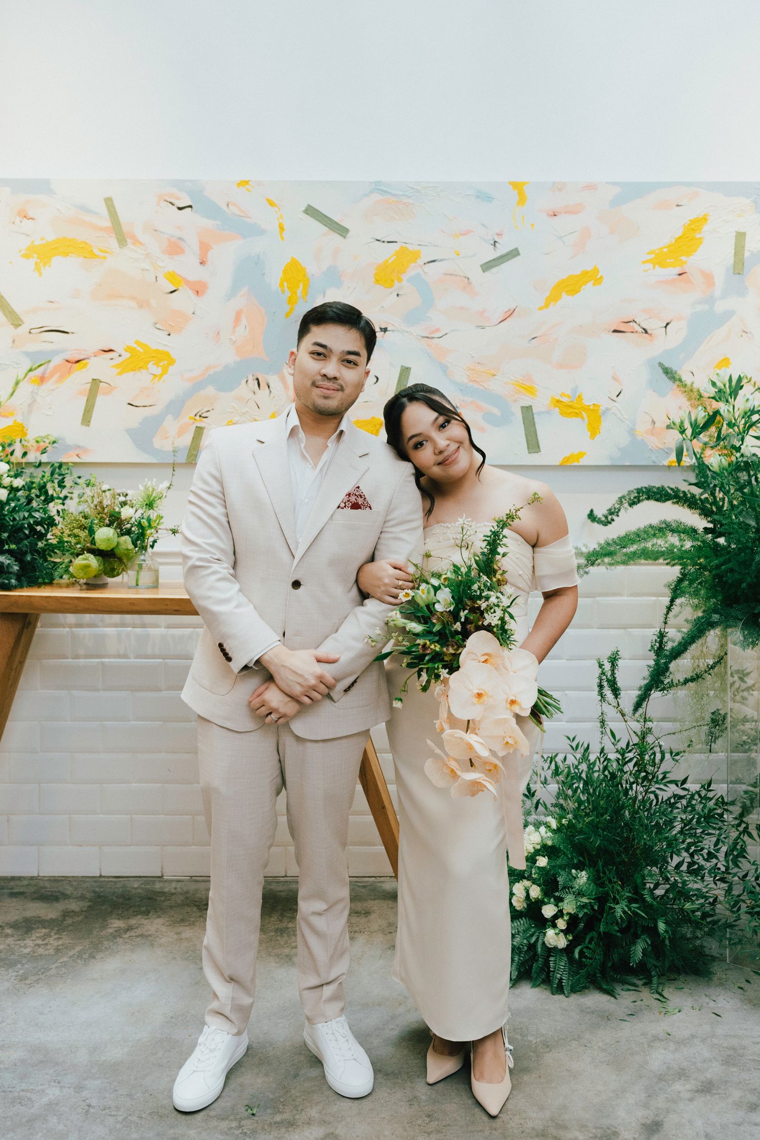 Bride and groom in matching cream outfits posing before colorful abstract art at wedding ceremony.