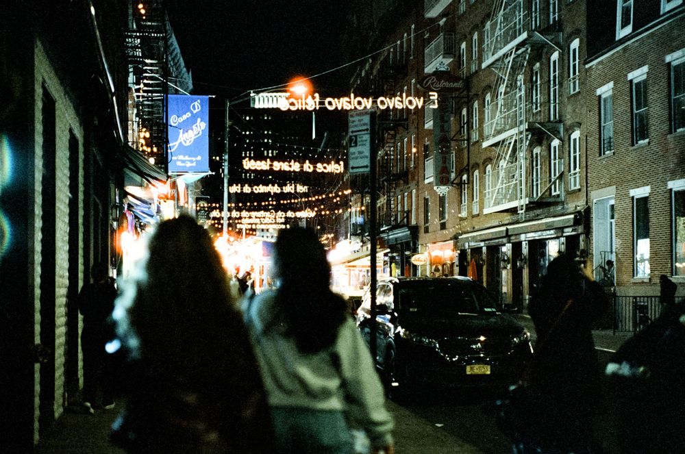 City street at night with neon signs and silhouettes of people walking.
