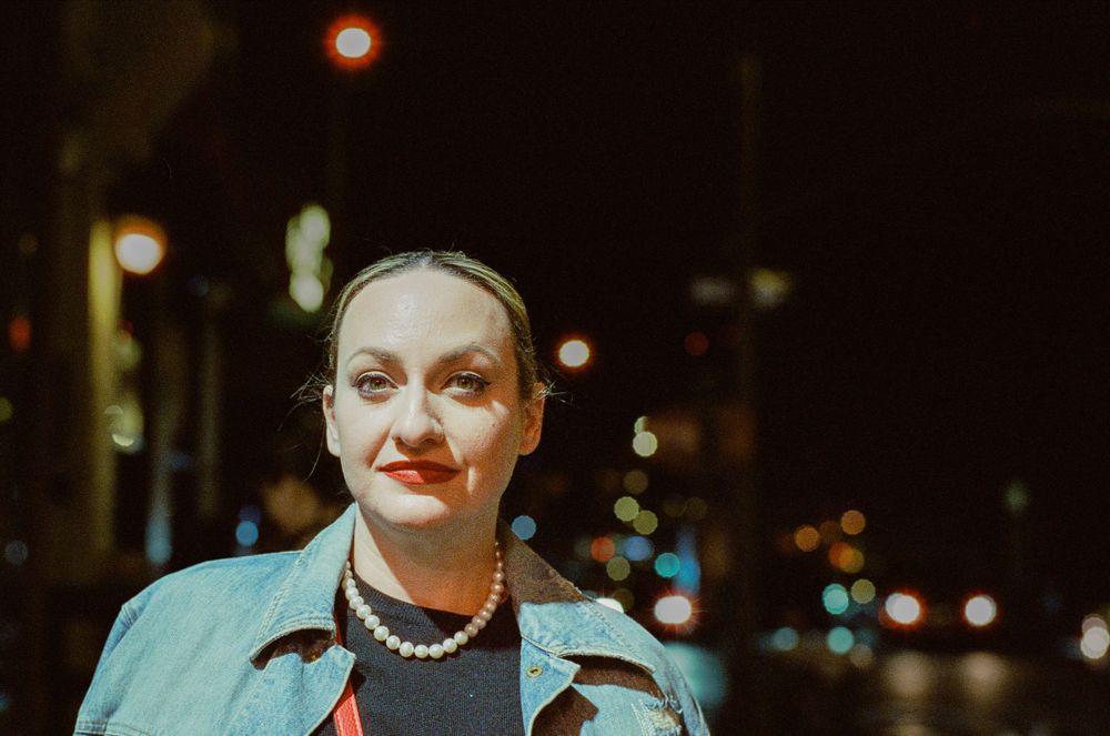 Person in turquoise jacket with pearl necklace photographed at night against city lights.