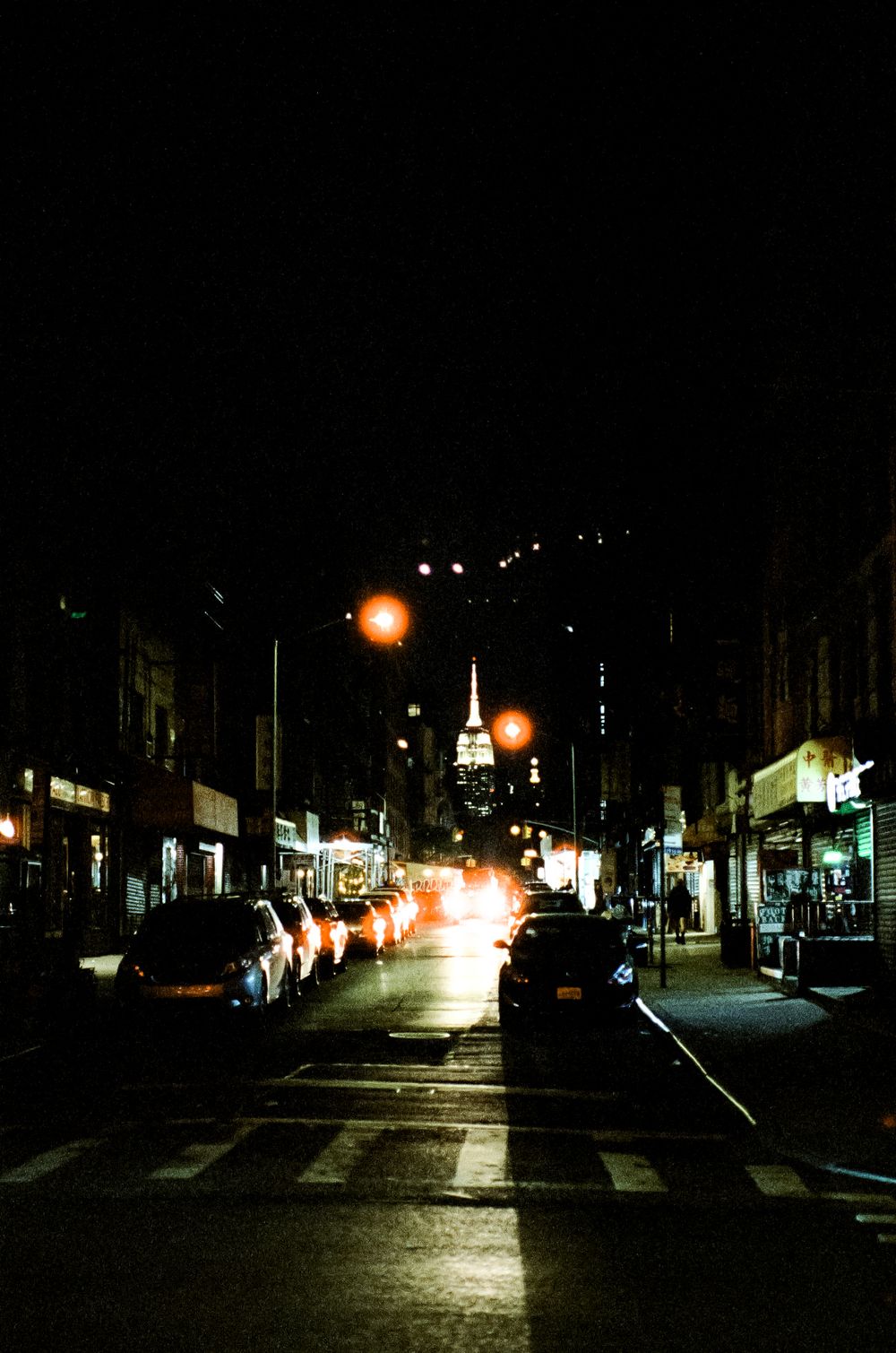 Dark urban street at night with cars parked along both sides and glowing streetlights.