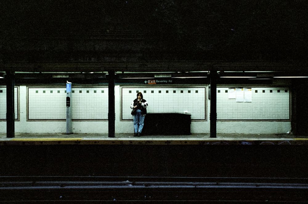 Lone figure waiting on empty subway platform with fluorescent lighting.