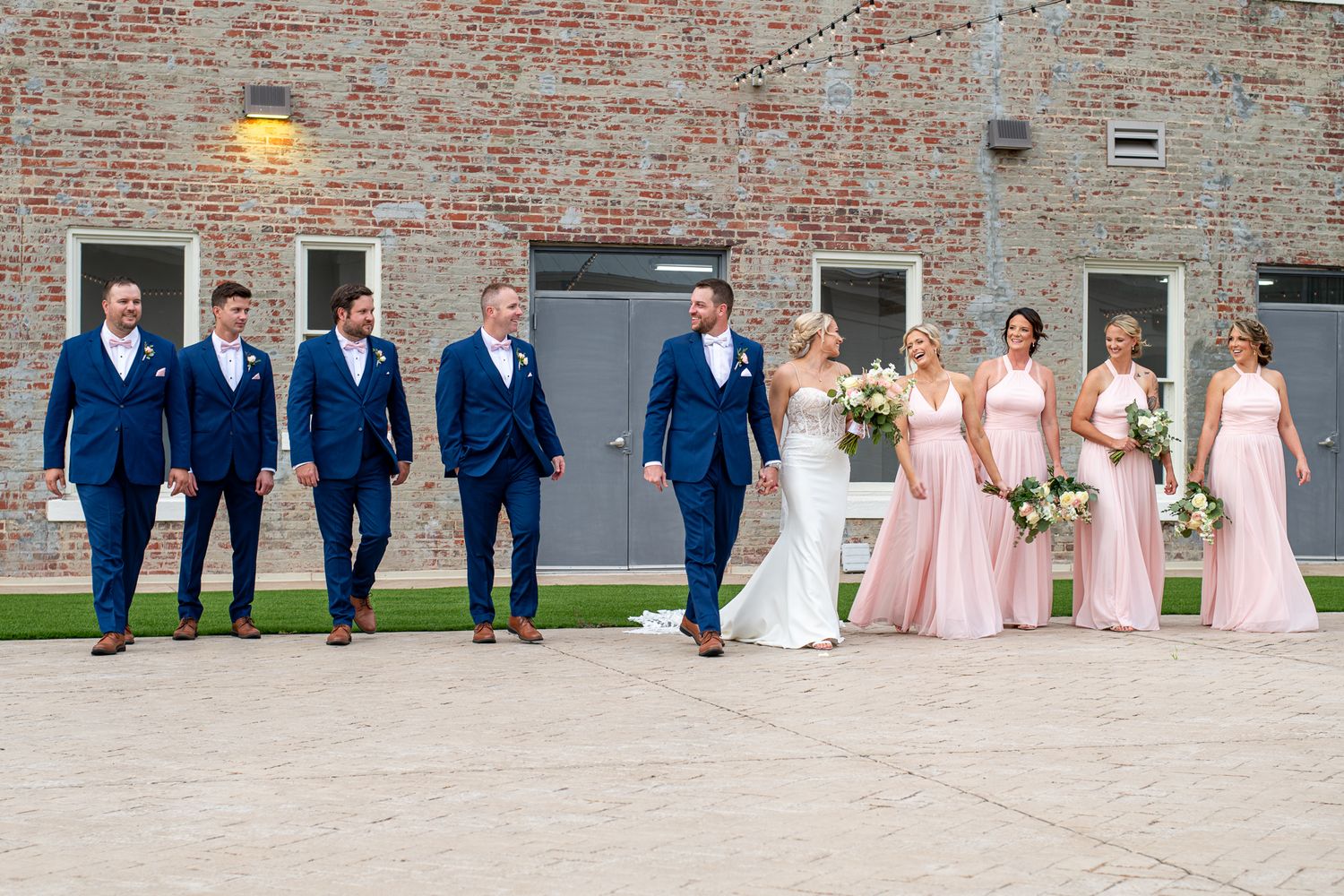 A wedding party in blue suits and pink dresses posed against a brick wall exterior.