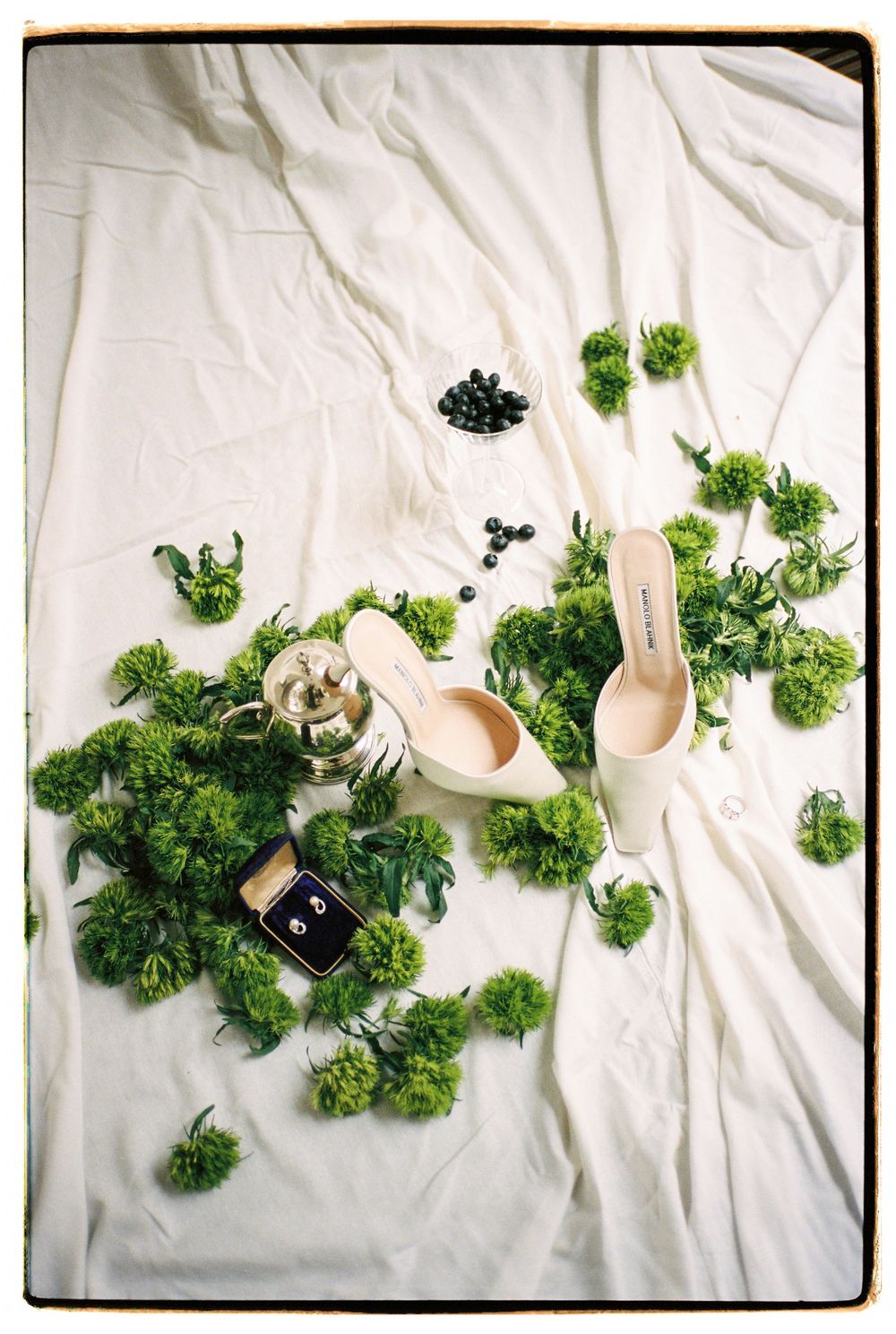 Wedding accessories and shoes artfully arranged with green florals on white fabric.