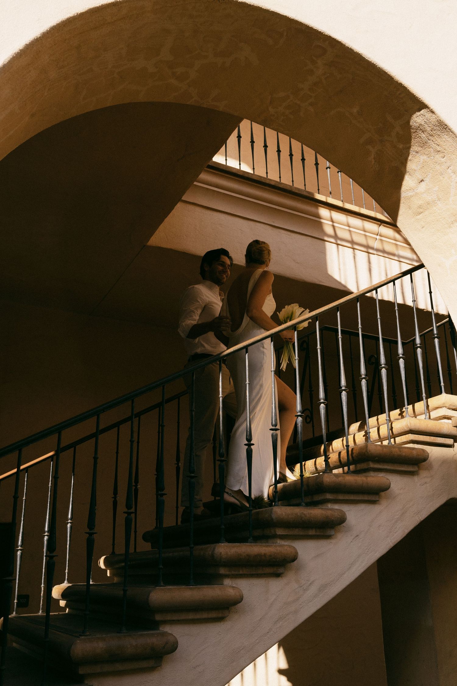 Two figures stand on a curved staircase under an arched ceiling with dramatic lighting.
