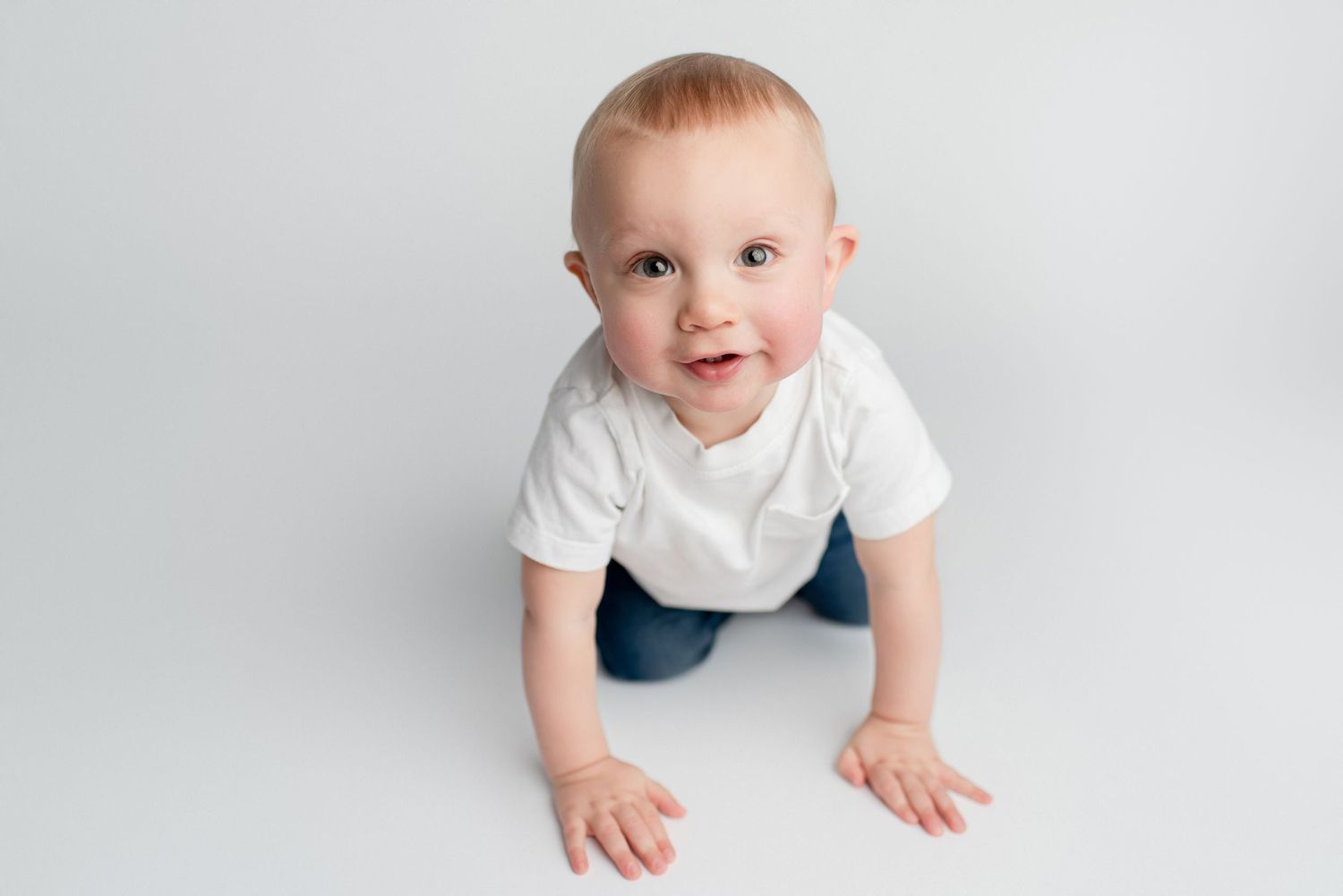 An adorable baby boy in a white onesie crawls toward the camera against a clean white background.