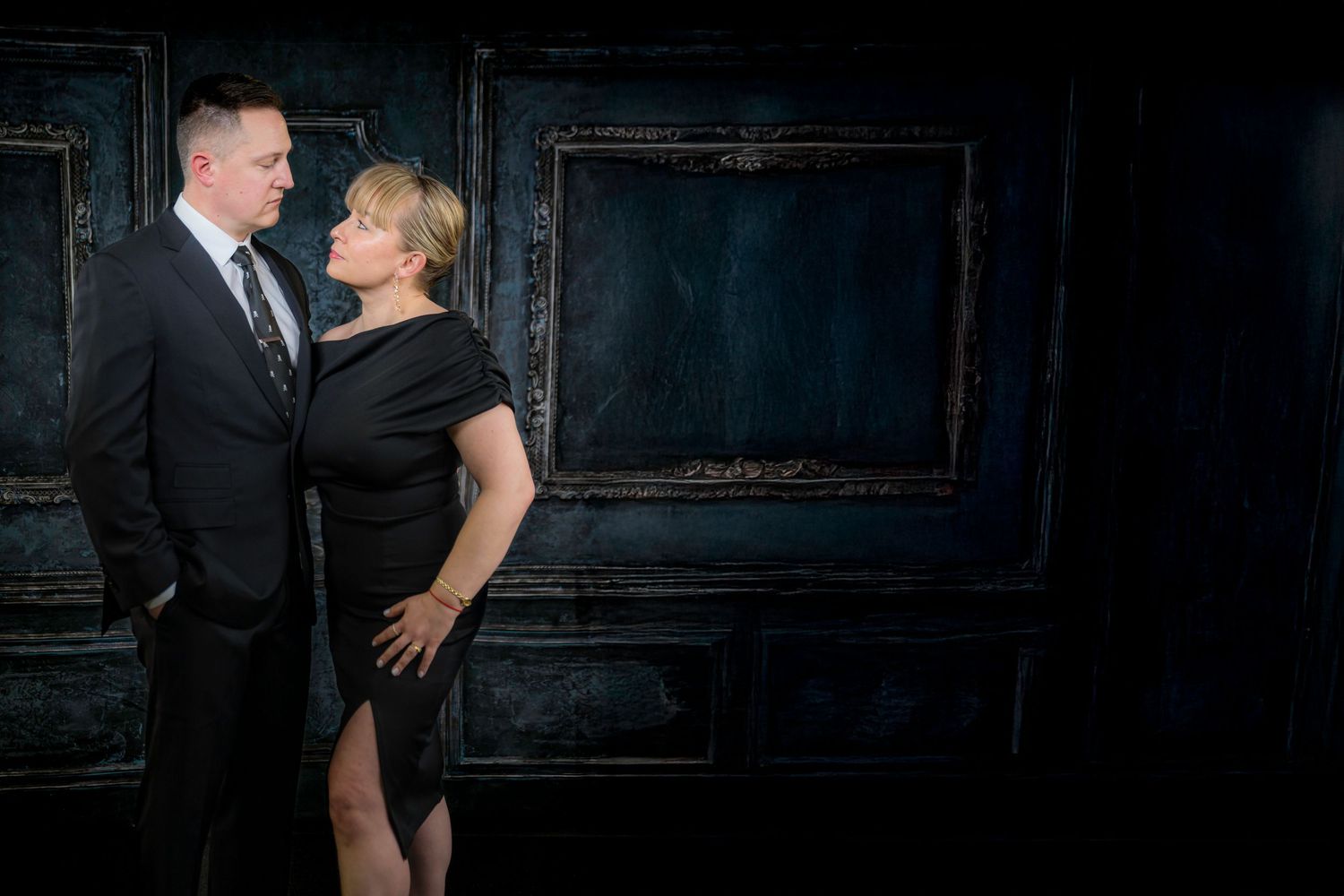 A romantic couple in elegant attire poses against a dark blue wall for formal portrait photographs.