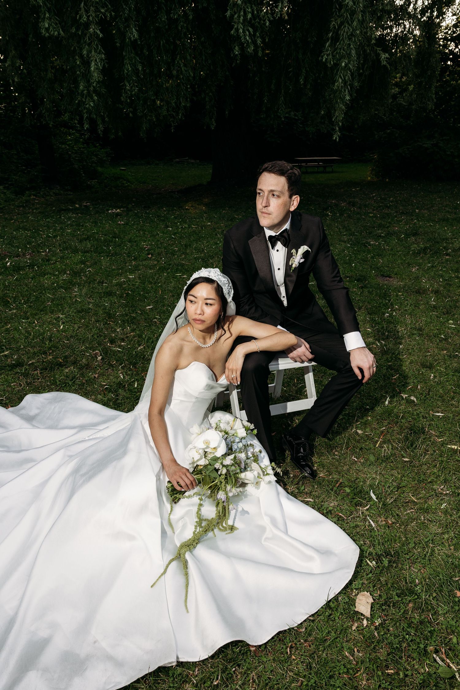 A bride in a white gown and groom in a black tuxedo pose together under weeping willow trees in a garden setting.