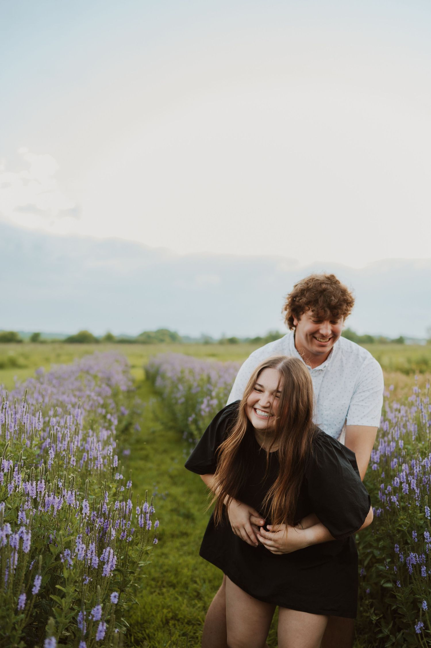 A couple shares joyful moments together while surrounded by blooming purple wildflowers in a sunny meadow.