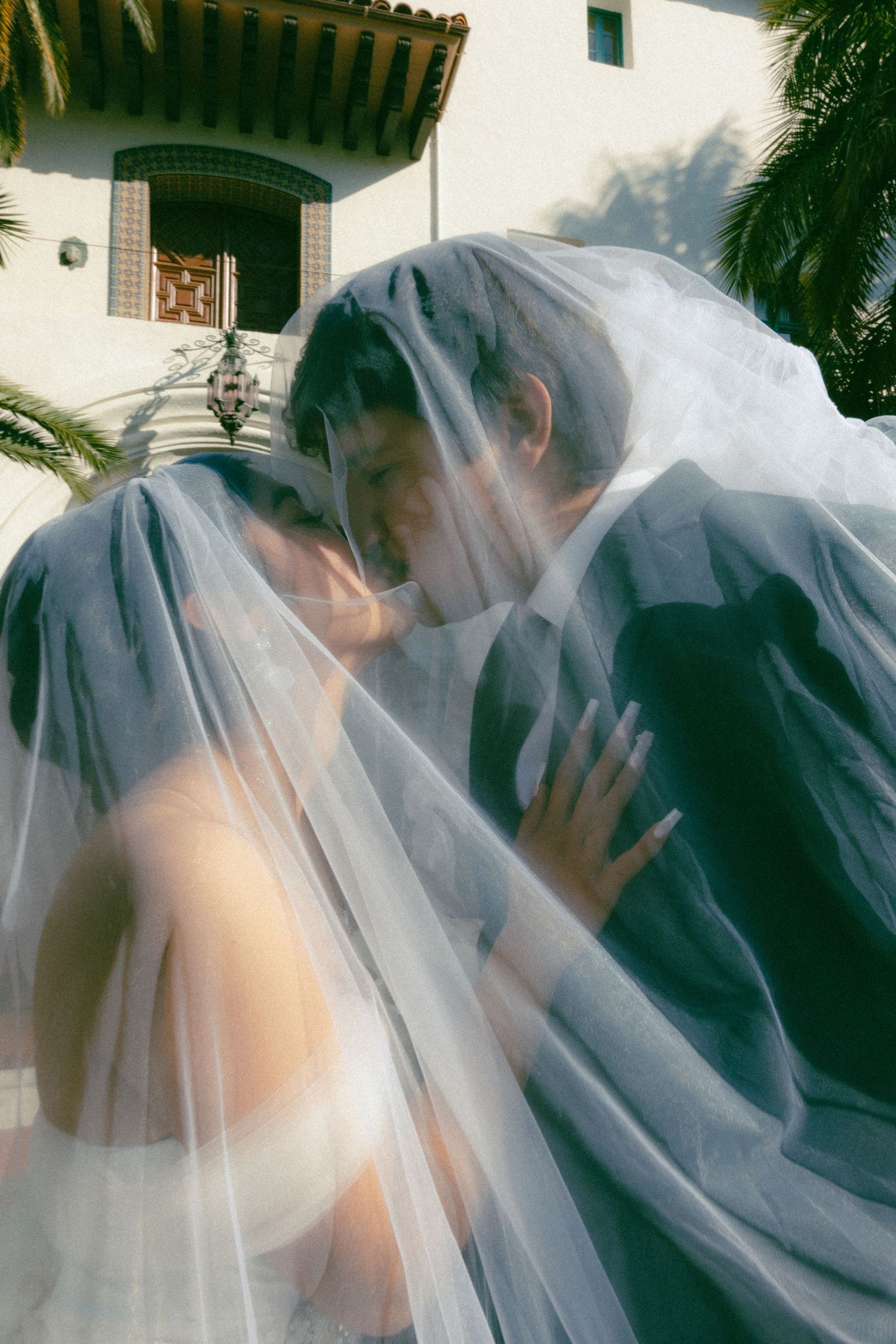 A romantic wedding photo under a sheer veil with Spanish architecture and palm trees in the background.