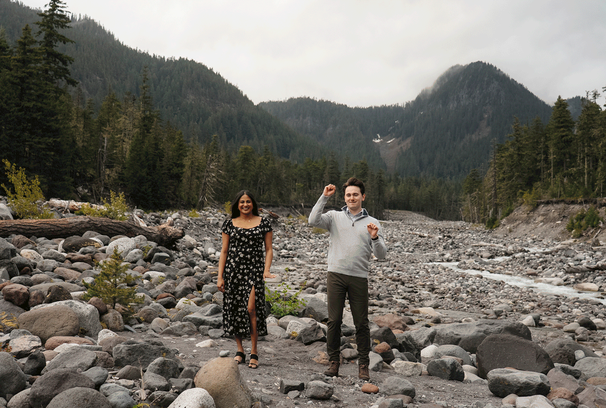 Two people stand on rocky riverbed surrounded by forested mountains in Pacific Northwest wilderness landscape.
