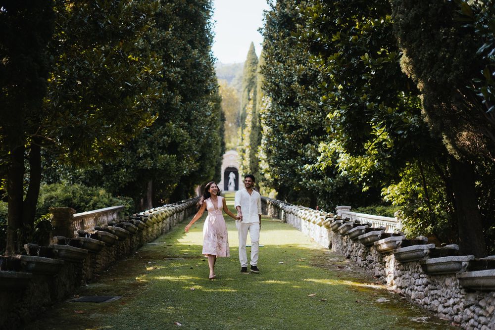 A couple in white outfits walks along a serene tree-lined pathway in a romantic garden setting.