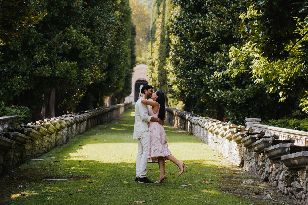 Couple in formal attire walks hand in hand along a stone wall path through lush garden scenery on their wedding day.