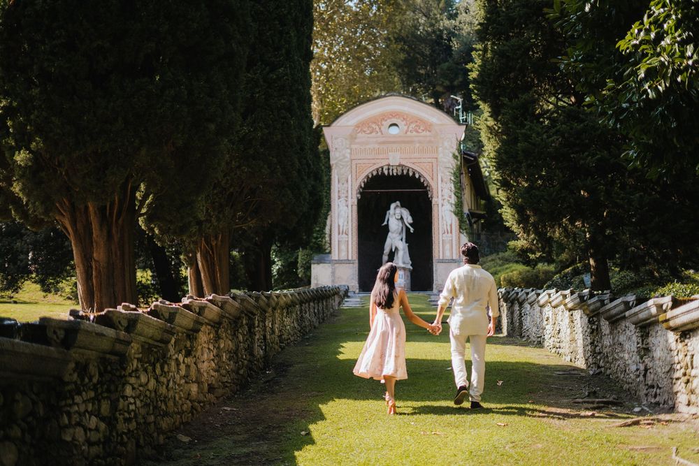 A couple walks together in a beautiful garden setting while wearing formal white attire on a sunny afternoon.