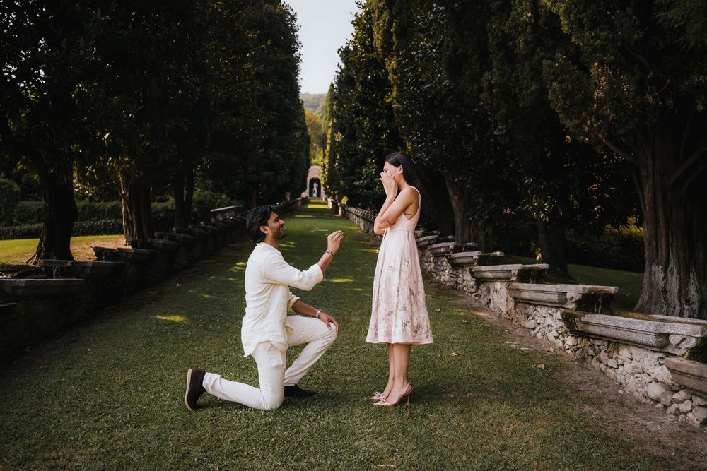 A romantic proposal moment captured in a lush garden setting with stone walls and cypress trees.