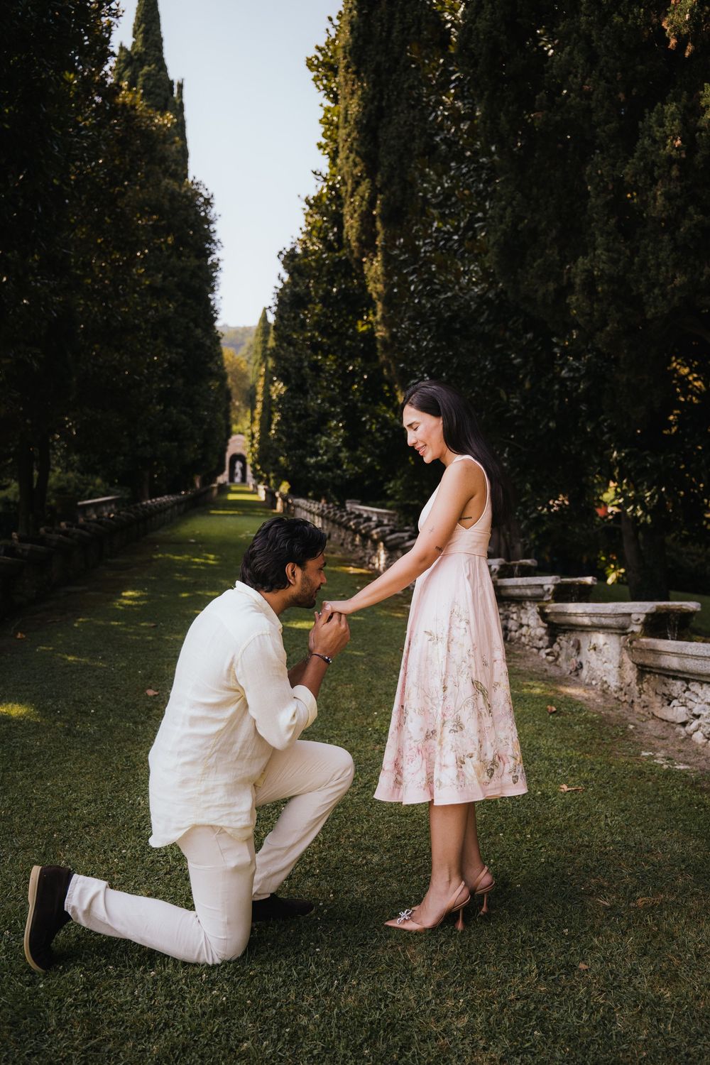 Romantic marriage proposal in a garden path lined with tall cypress trees and stone balustrades.