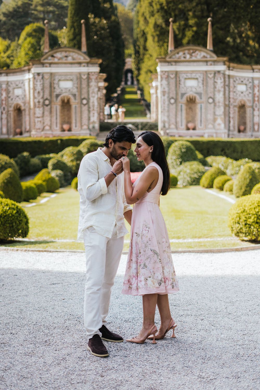 A romantic couple in coordinated white and pink outfits share an intimate moment in front of ornate garden architecture.