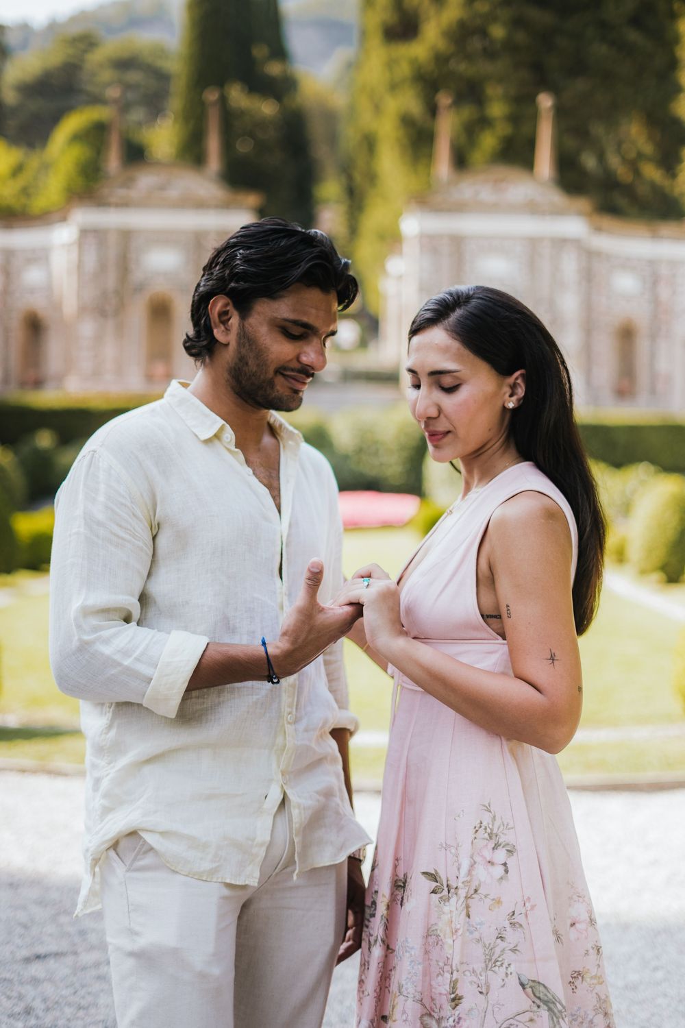 A romantic couple in light colored outfits share an intimate moment together in a garden with ornate architecture.