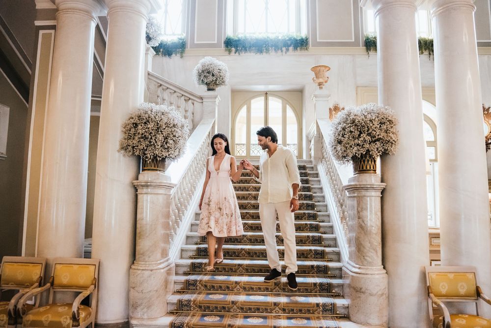 A couple descends an elegant marble staircase framed by large floral arrangements and ornate columns in a grand venue.