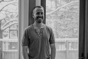A person wearing a gray v-neck t-shirt smiles warmly by a window in a black and white portrait.