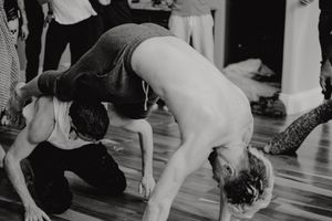 Two dancers perform an acrobatic dance move during a studio practice session in black and white.
