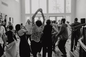 A black and white photo of a lively dance gathering with people moving in a bright studio space.