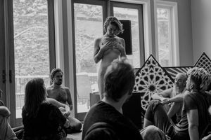 Standing performer addresses seated audience members near large windows with geometric patterns.