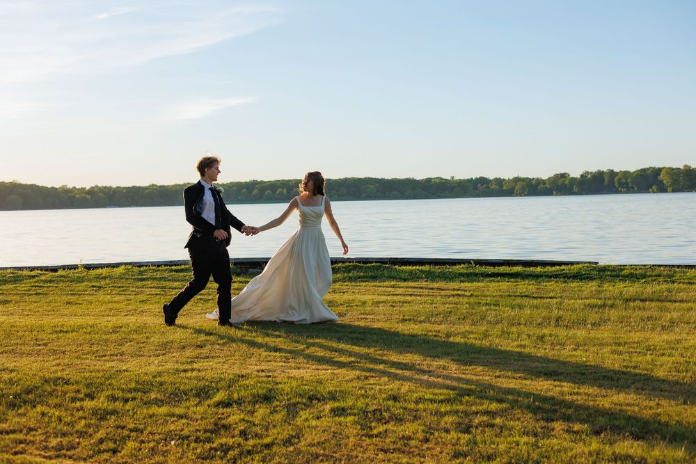 Bride and groom holding hands running on green lawn by a lake on a sunny wedding day.