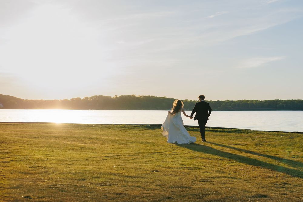 Bride and groom walking hand in hand toward a serene lakeside sunset on lush green grass.