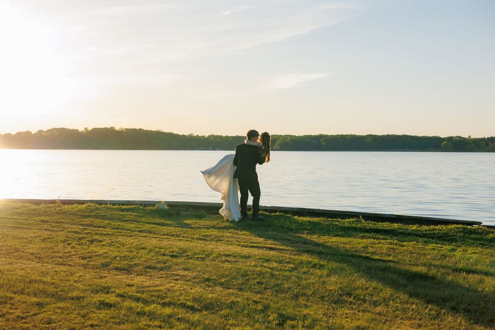 Couple embracing by a lakeside at golden hour, bride in flowing white gown, lush green grass and tree-lined horizon.