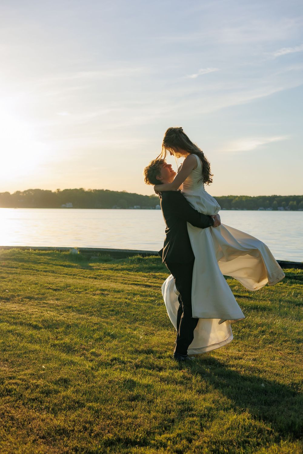 Groom lifts bride in white gown at sunset by waterfront, sharing romantic moment on lush green lawn.