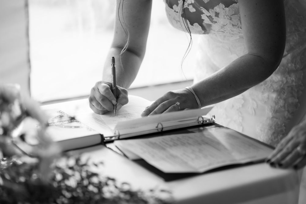 Bride in lace wedding dress signing marriage certificate at elegant ceremony table in black and white photo.