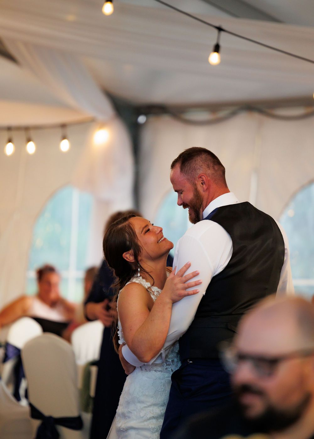 Bride and groom share their first dance under string lights in a white draped wedding reception tent.