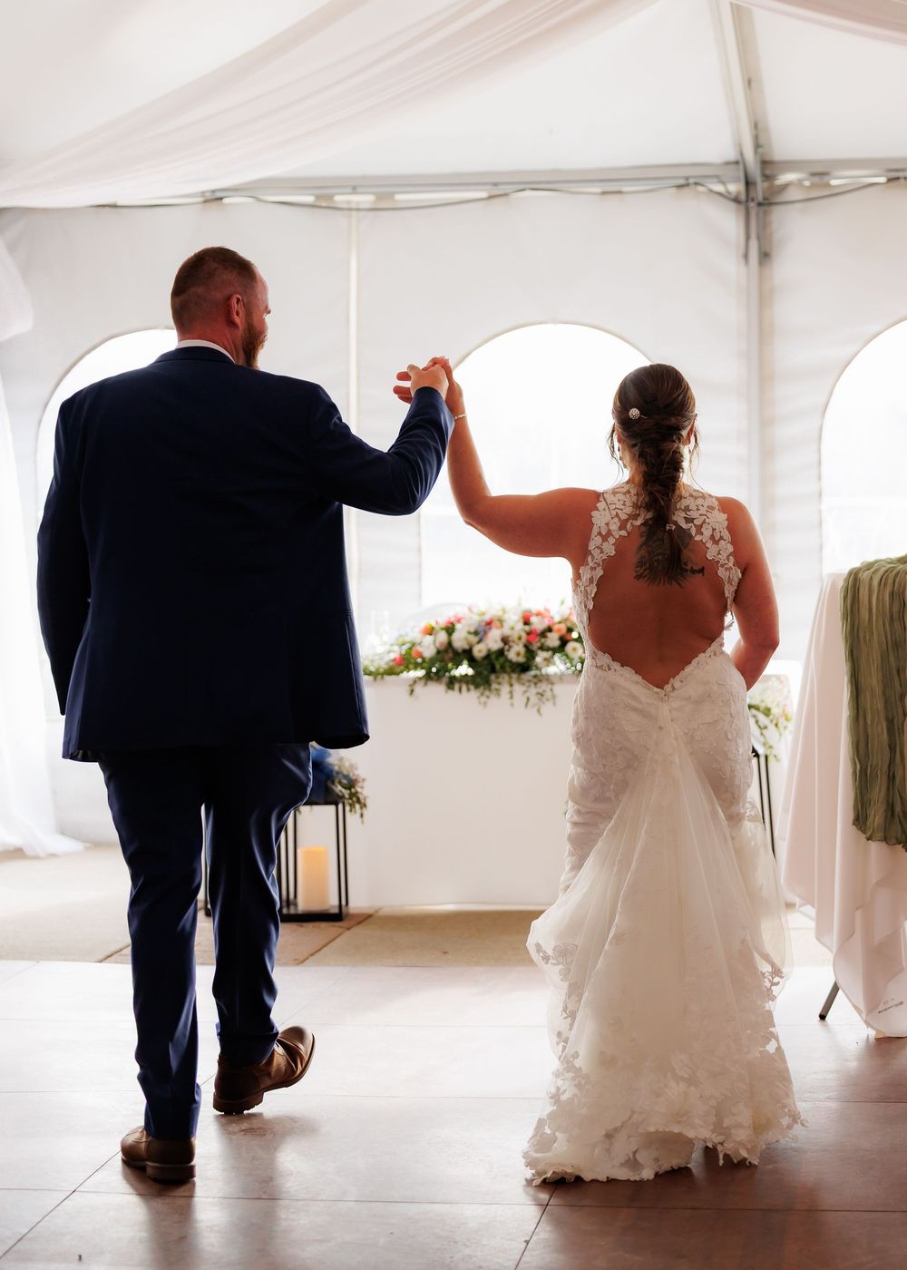Bride and groom dancing hand in hand under a white wedding tent, bride wearing a lace backless gown.